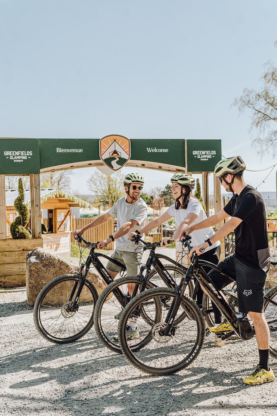 Three people with bicycles at a wooden archway with 'Greenfields Glamping' signage.
