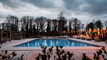 A rectangular swimming pool with a wooden deck, surrounded by lounge chairs and umbrellas, and trees in the background.