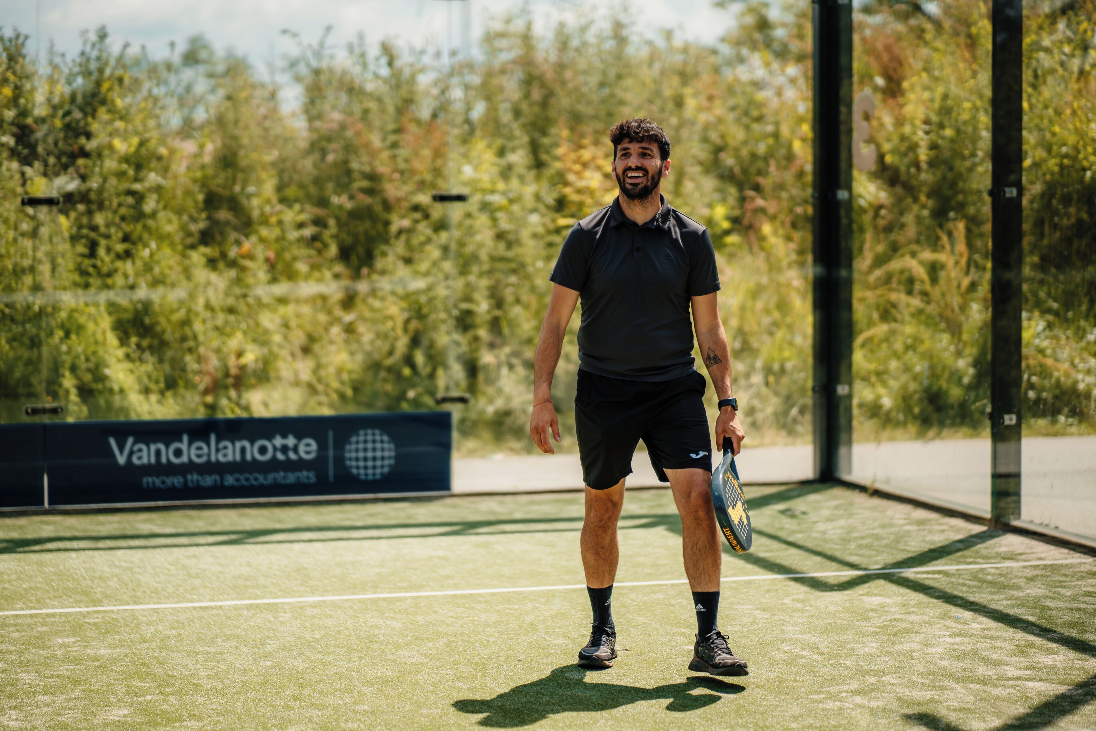 En tennisbana med en person som håller i ett racket, en skylt med texten ”Vandelanotte – mer än bara revisorer” och grönska i bakgrunden.