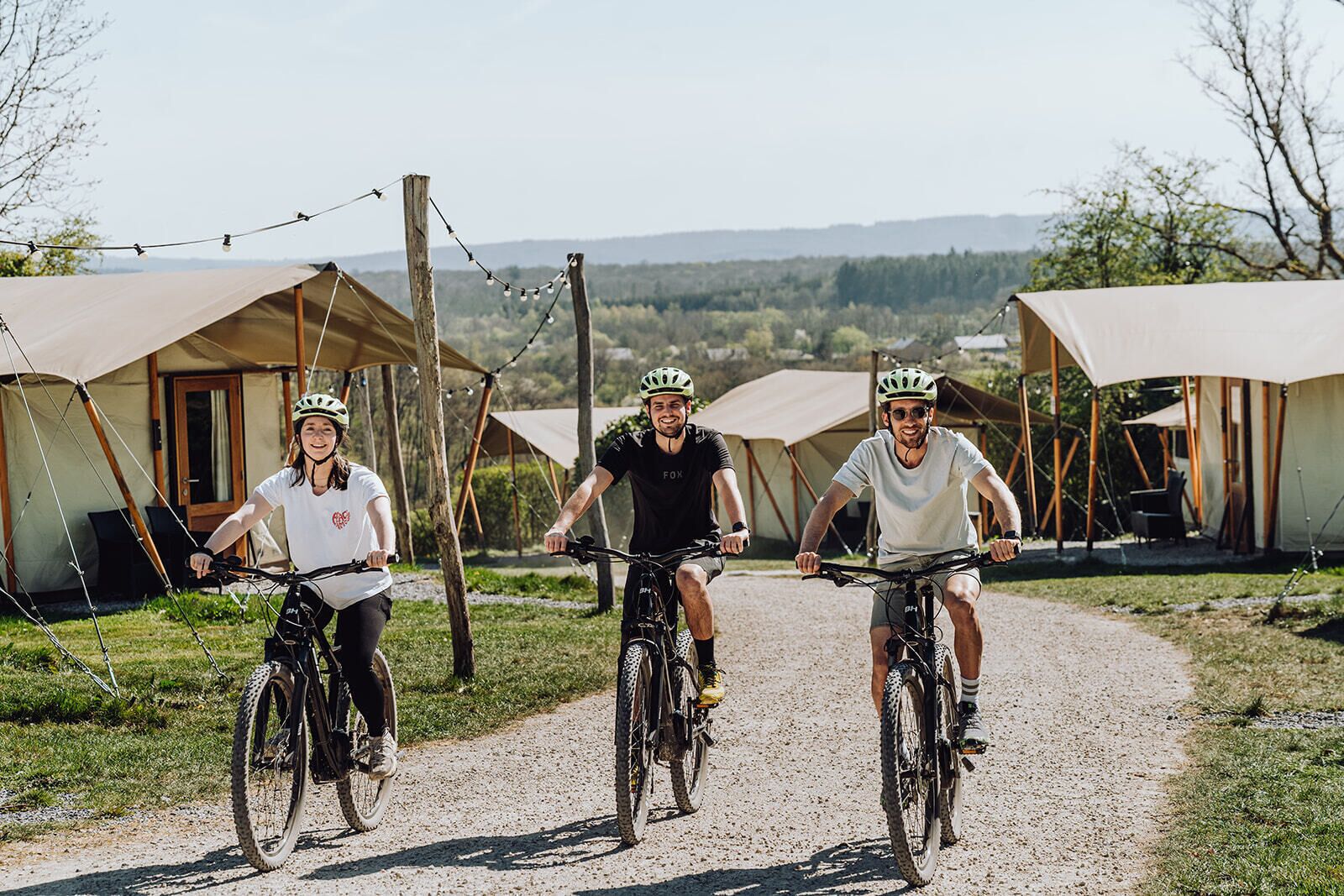Tres personas en bicicletas sobre un camino de grava con tiendas de campaña al fondo.