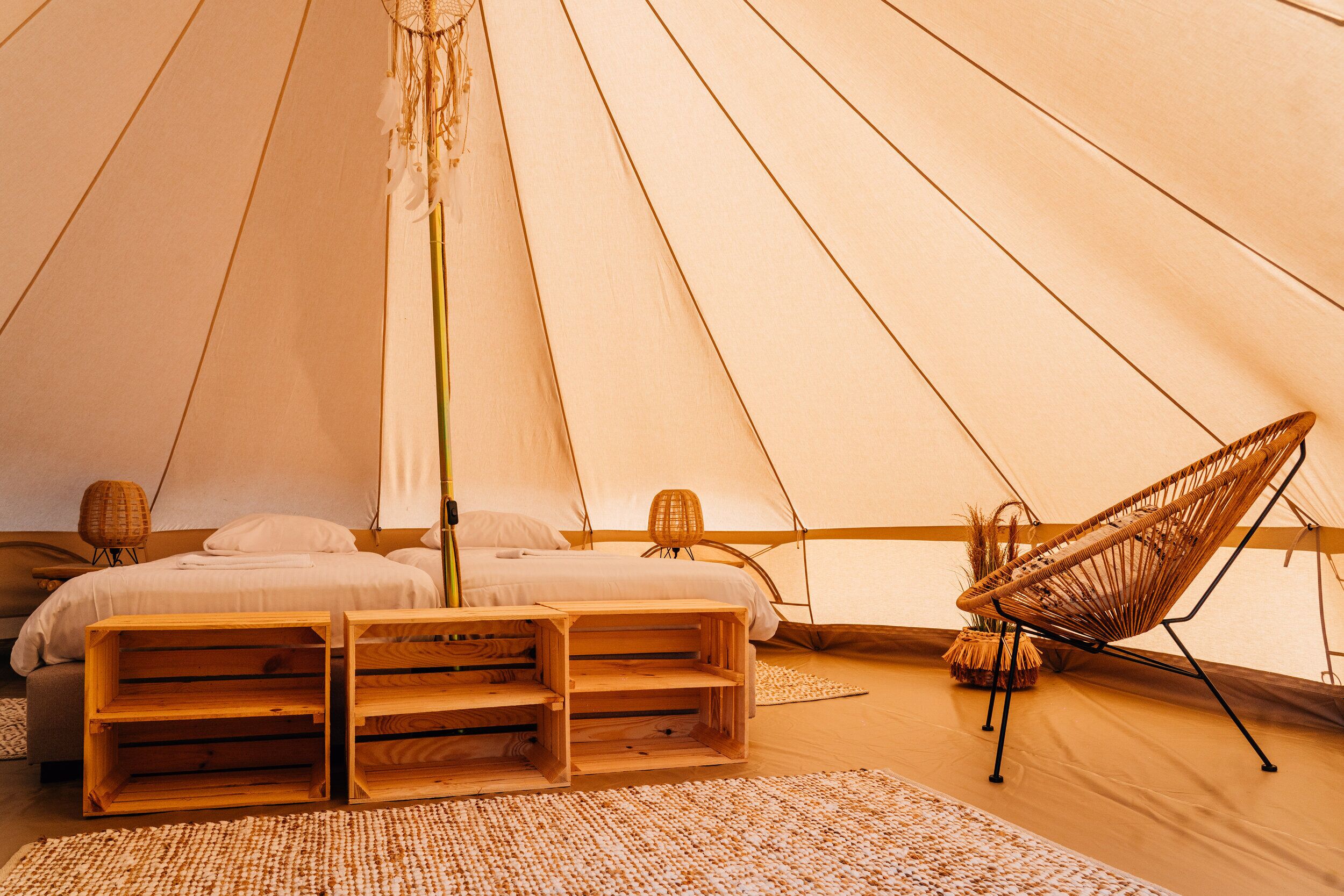 Uma tenda com uma cama, caixas de madeira, uma cadeira de fibra natural e uma luminária de fibra natural.