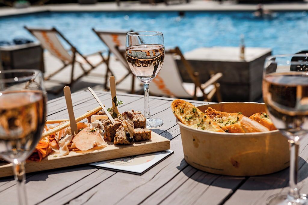 Une table en bois installée au bord de la piscine, avec un panier de pain et des verres de vin.