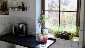 Une cuisine équipée d’une machine à café, d’un panier rempli de légumes, d’une plante en pot et d’une fenêtre donnant sur des arbres.