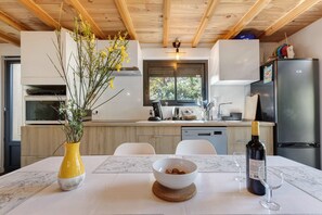 A modern kitchen with a dining table, a vase with flowers, a bowl of nuts, a wine bottle, and glasses. - Chalet "Seafront" with Private Terrace, Garden and Wi-Fi (Le Verdon-sur-Mer)