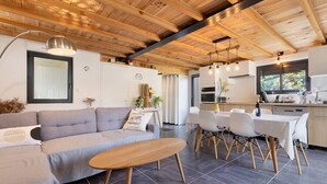 A modern interior with a wooden ceiling, a dining area with white chairs, a grey sofa, and a wooden coffee table. - Chalet "Seafront" with Private Terrace, Garden and Wi-Fi (Le Verdon-sur-Mer)