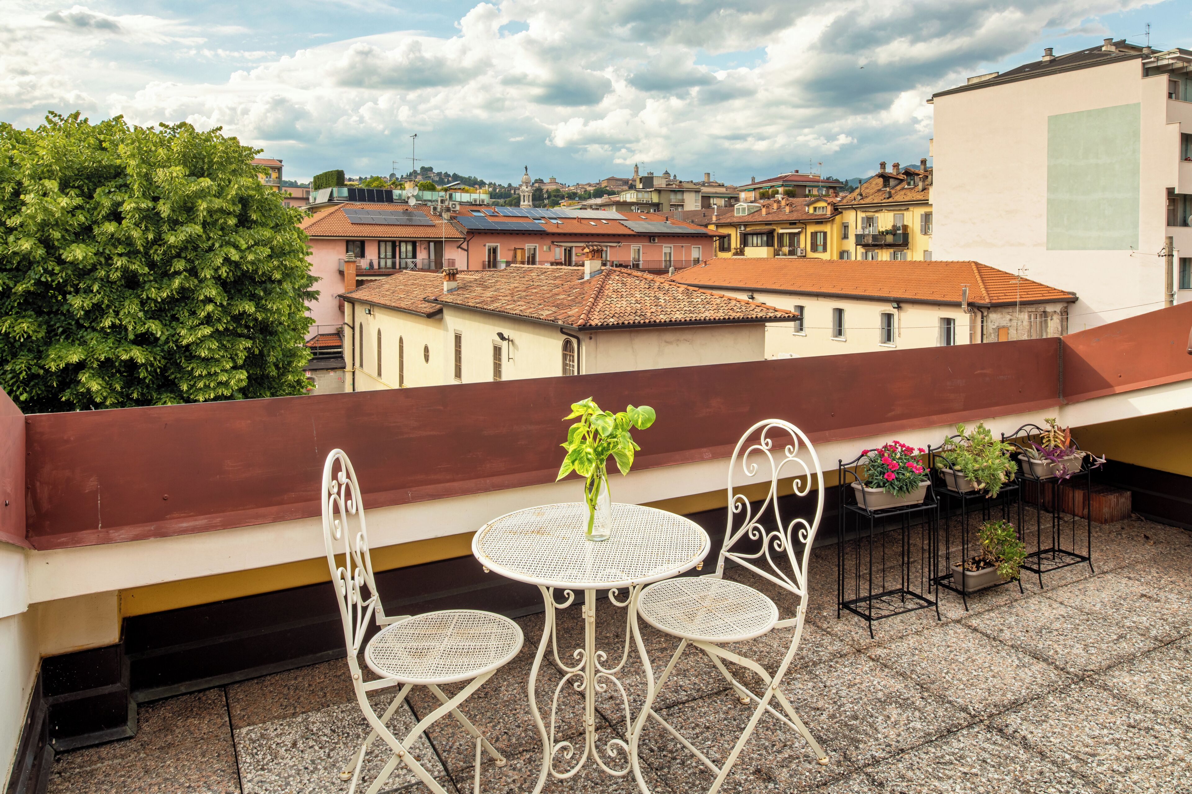 A rooftop terrace with a table and two chairs, overlooking a cityscape with terracotta-roofed buildings.