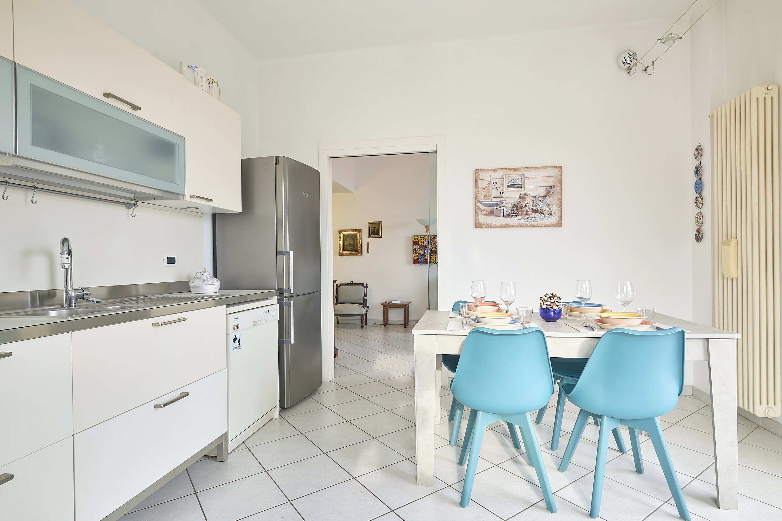 Une cuisine moderne avec des meubles blancs, un réfrigérateur en acier inoxydable et un coin repas comprenant une table blanche et des chaises bleues.