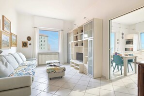 Un salon moderne avec un canapé, un pouf et un téléviseur posé sur un meuble. On y trouve également un coin repas avec des chaises et une table, et une cuisine visible à travers une porte ouverte.