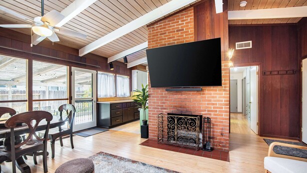 A spacious living area with a fireplace, a flat-screen TV mounted on a brick wall, wooden flooring, and a ceiling fan. - Spacious 4BR Backyard Oasis with BBQ Near UC Davis (Davis)