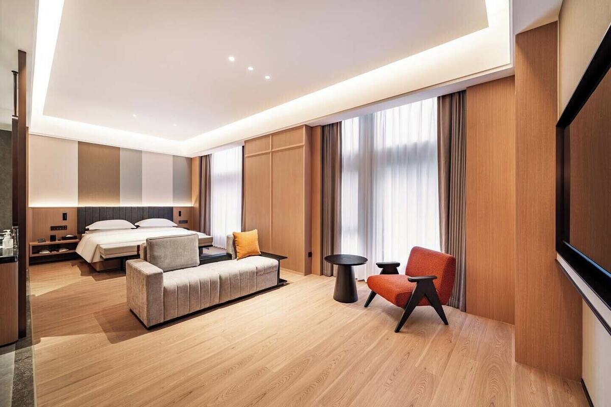 A modern hotel room with a bed, sofa, two chairs, a TV, and a large window with curtains.