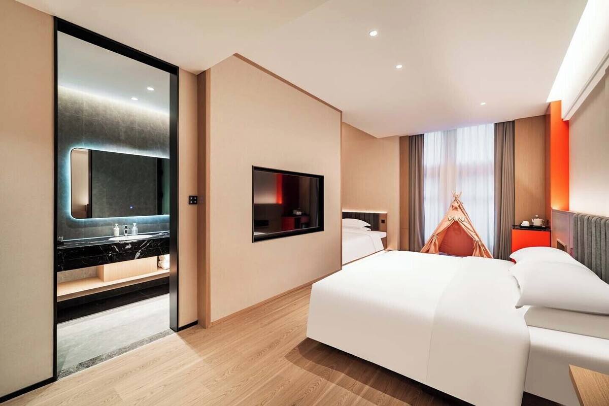 A hotel room with a bed, a TV mounted on the wall, a mirror, a wooden floor, and a window with curtains.