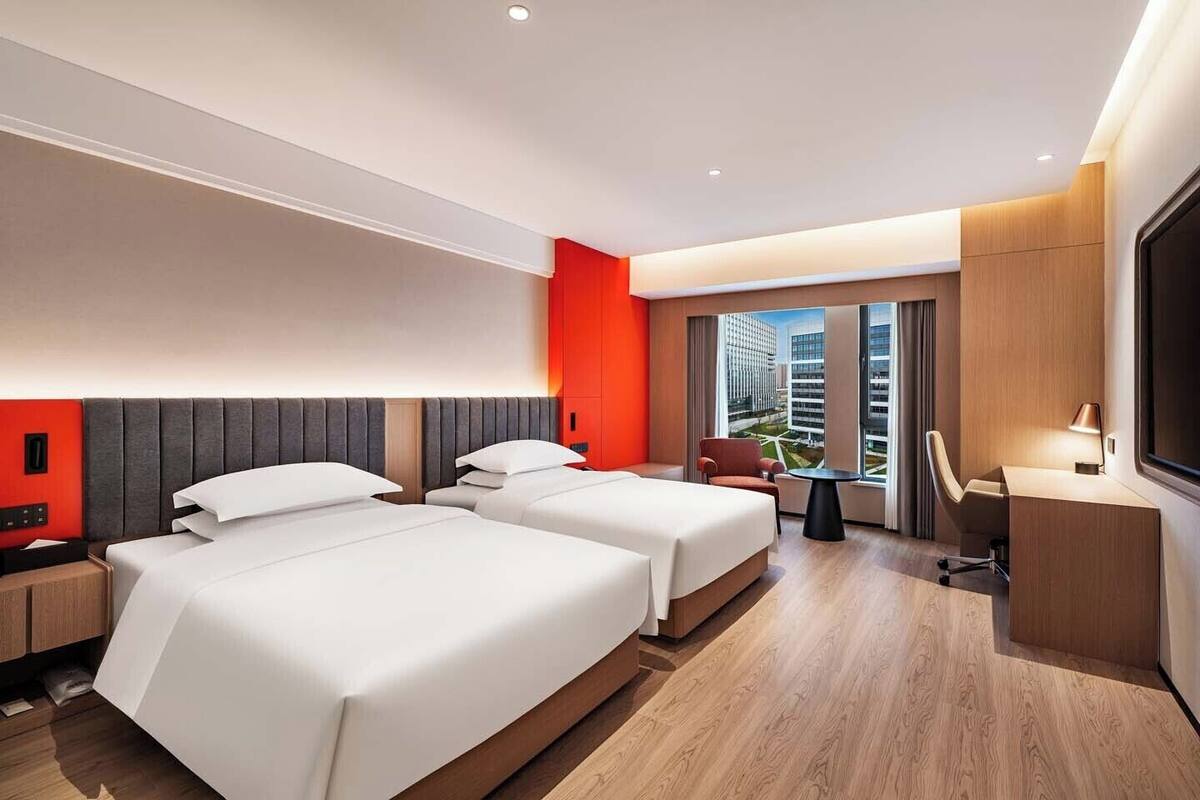 A hotel room with two beds, a desk, a chair, and a large window with a city view.