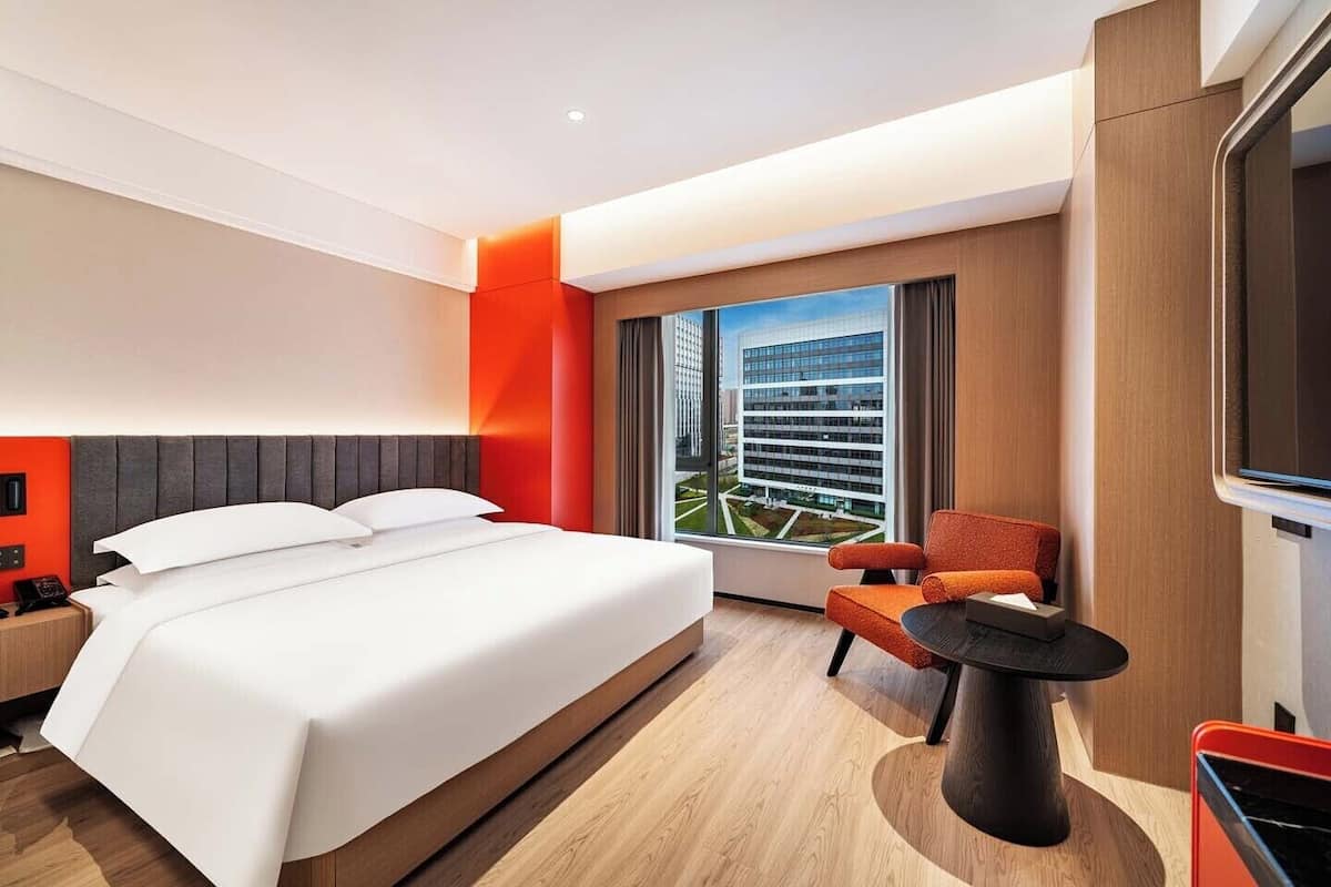 A modern hotel room with a large bed, a seating area with a table, and a view of a cityscape through a large window.