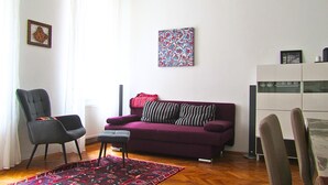 Ein Wohnzimmer mit einem violetten Sofa, einem schwarzen Sessel, einem kleinen Tisch, einem gemusterten Teppich und einer Wand mit einem gerahmten Bild.