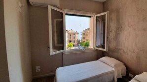 Ein kleines Zimmer mit einem Einzelbett, einem Fenster mit Blick auf Gebäude und einer schlichten Wand.