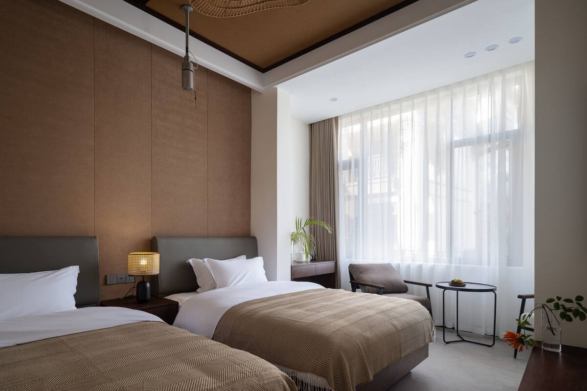 Ein Hotelzimmer mit zwei Betten, einem kleinen Tisch, einem Sessel, einer Lampe und einem großen Fenster mit durchsichtigen Vorhängen.