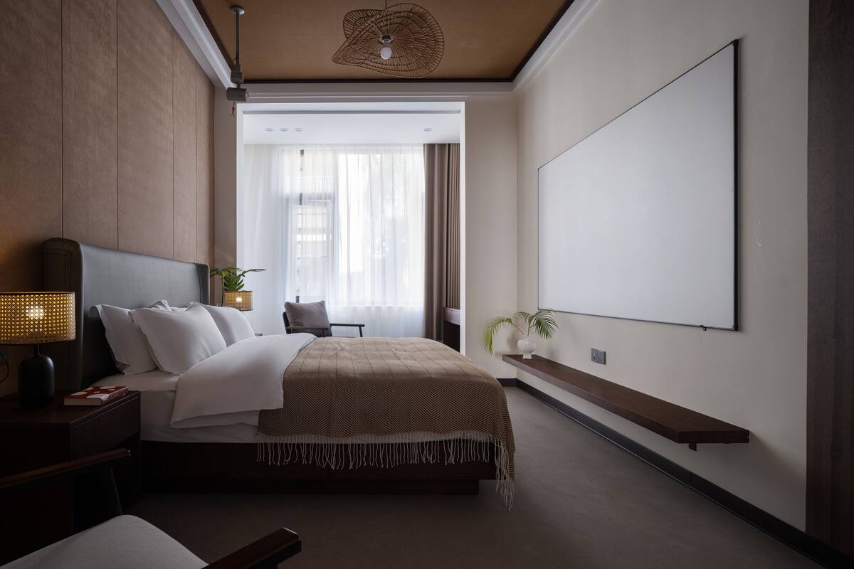 Ein modernes Hotelzimmer mit einem großen Bett, einem Schreibtisch, einem Sessel, einem Nachttisch, einer Lampe, einer Pflanze, einem an der Wand montierten Fernseher und einem Fenster mit durchsichtigen Vorhängen.