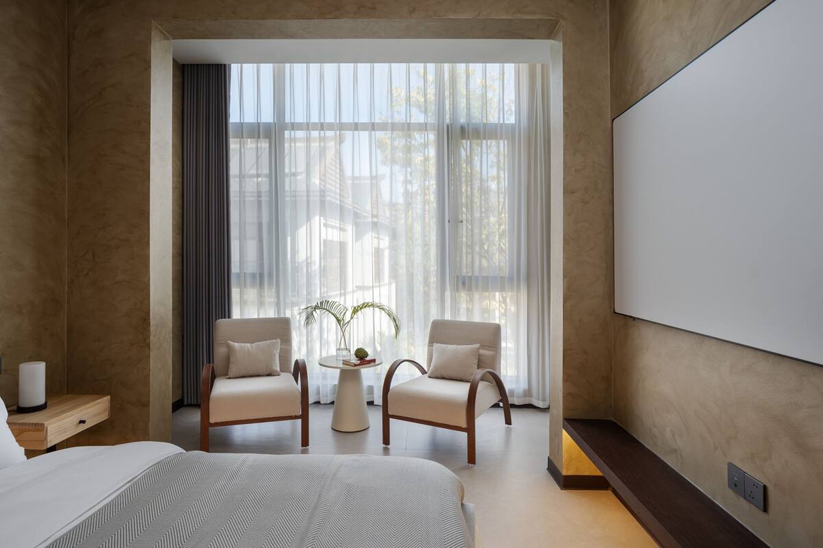 Ein Hotelzimmer mit einem Bett, zwei Sesseln, einem kleinen Tisch und einem großen Fenster mit durchsichtigen Vorhängen.