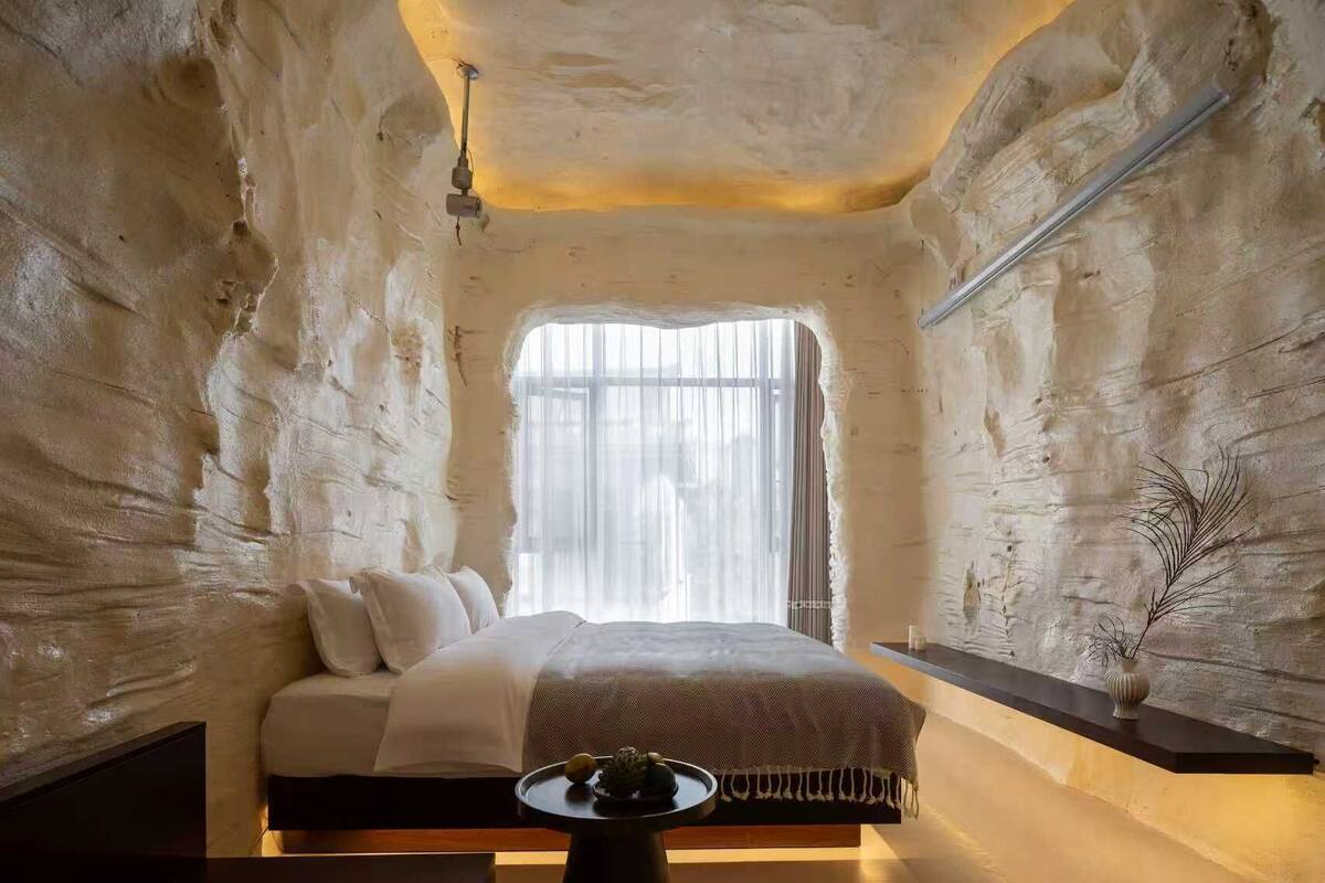 Ein Zimmer mit Bett, Fenster, einer Wandablage und einem Tisch mit Pflanze.