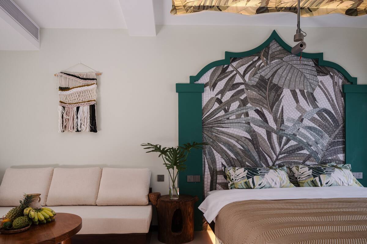Ein Schlafzimmer mit Bett, einer Couch, einem kleinen Holztisch mit Obst, einem Wandbehang aus Makramee und einer großen Wandmalerei mit Blättermotiv.