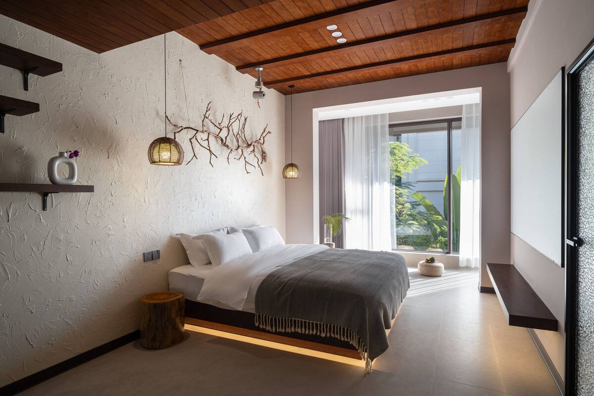 Ein modernes Schlafzimmer mit einem großen Bett, Holzbalkendecke, einer Zweig-Wanddekoration und einem großen Fenster mit Blick auf Grünflächen.