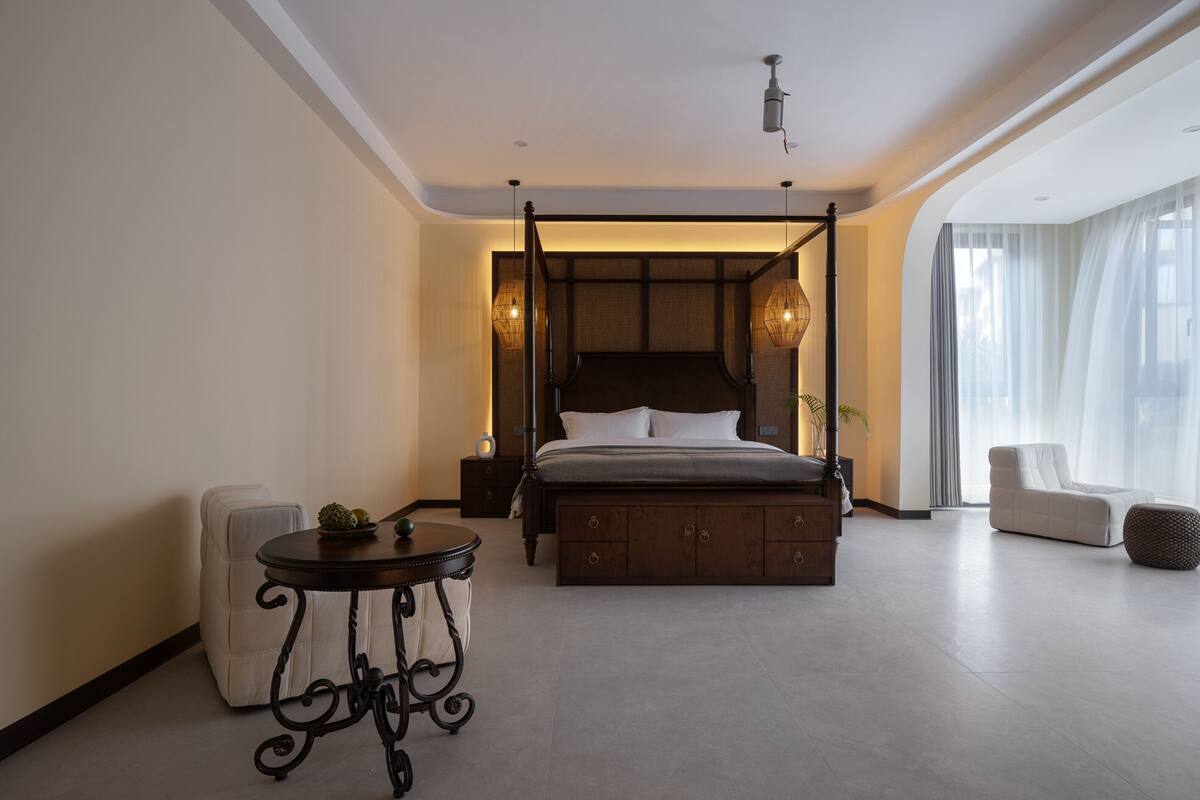 Ein modernes Schlafzimmer mit einem Himmelbett, einem runden Tisch mit Pflanze und einem großen Fenster mit transparenten Vorhängen.