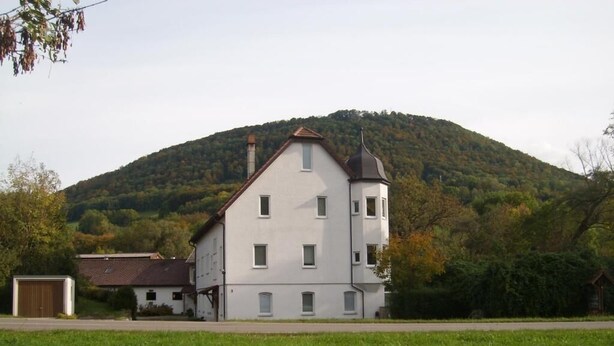 A two-story building with a symmetrical facade, a tower on the right, and a hill in the background.