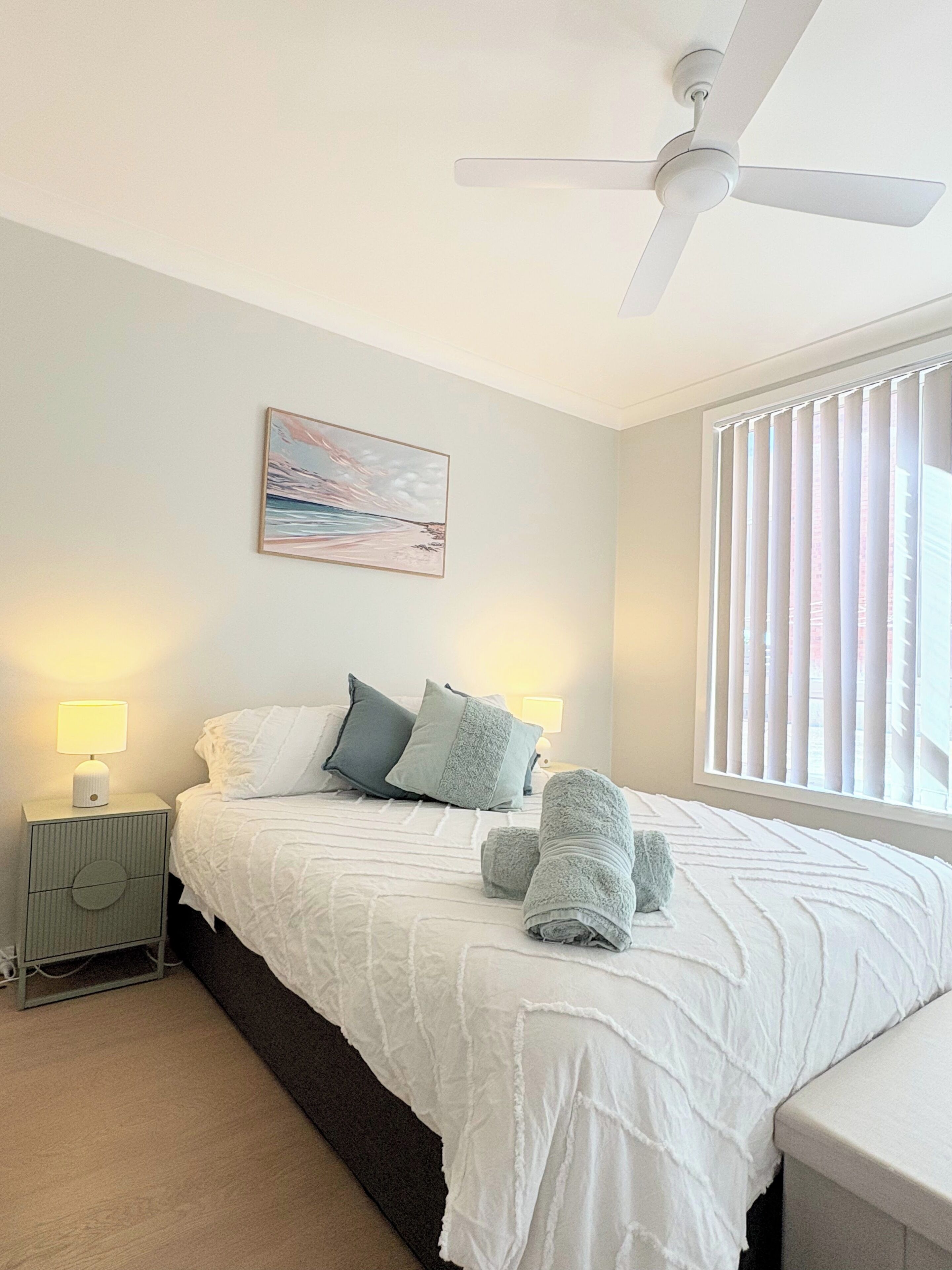 A bedroom with a bed, two bedside lamps, a ceiling fan, a window with blinds, and a painting on the wall.