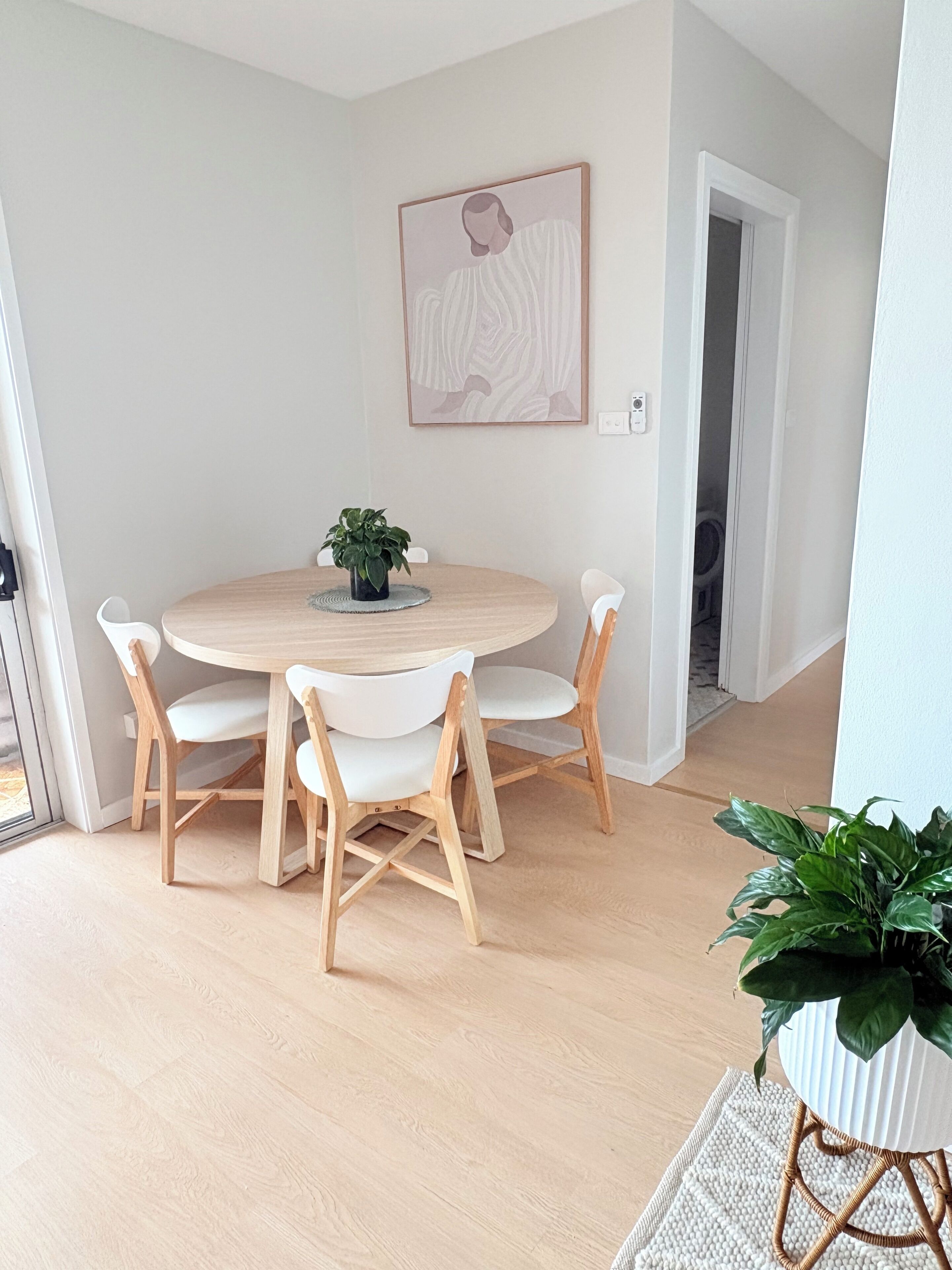 A dining area with a round table, four chairs, a potted plant, and a framed artwork on the wall.