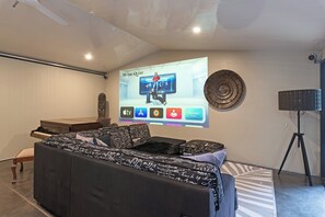 A modern living room with a large screen displaying a movie, a piano, a sofa, and a decorative wall piece. - Home among the Gum Trees - free wine (Tea Tree Gully)