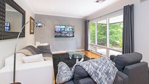 A modern living room with a large TV, comfortable seating, and a view through large windows. - Home among the Gum Trees - free wine (Tea Tree Gully)