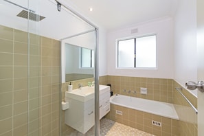 A modern bathroom with a glass shower enclosure, a white vanity with a mirror, a bathtub, and a window providing natural light.