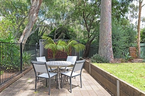 A patio with a round table and four chairs, surrounded by a black metal fence and lush greenery.