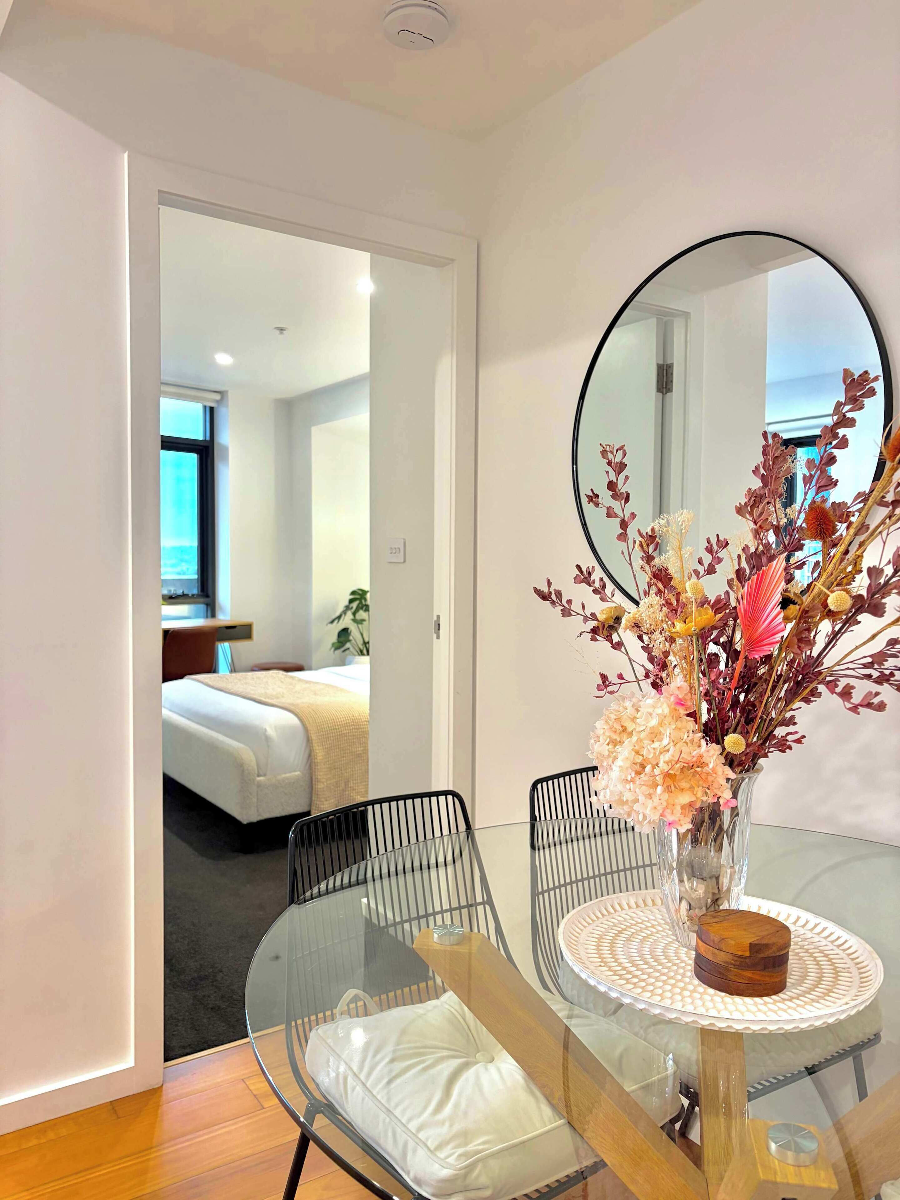 A hotel room with a bed, a bedside table, a mirror, and a glass table with chairs.