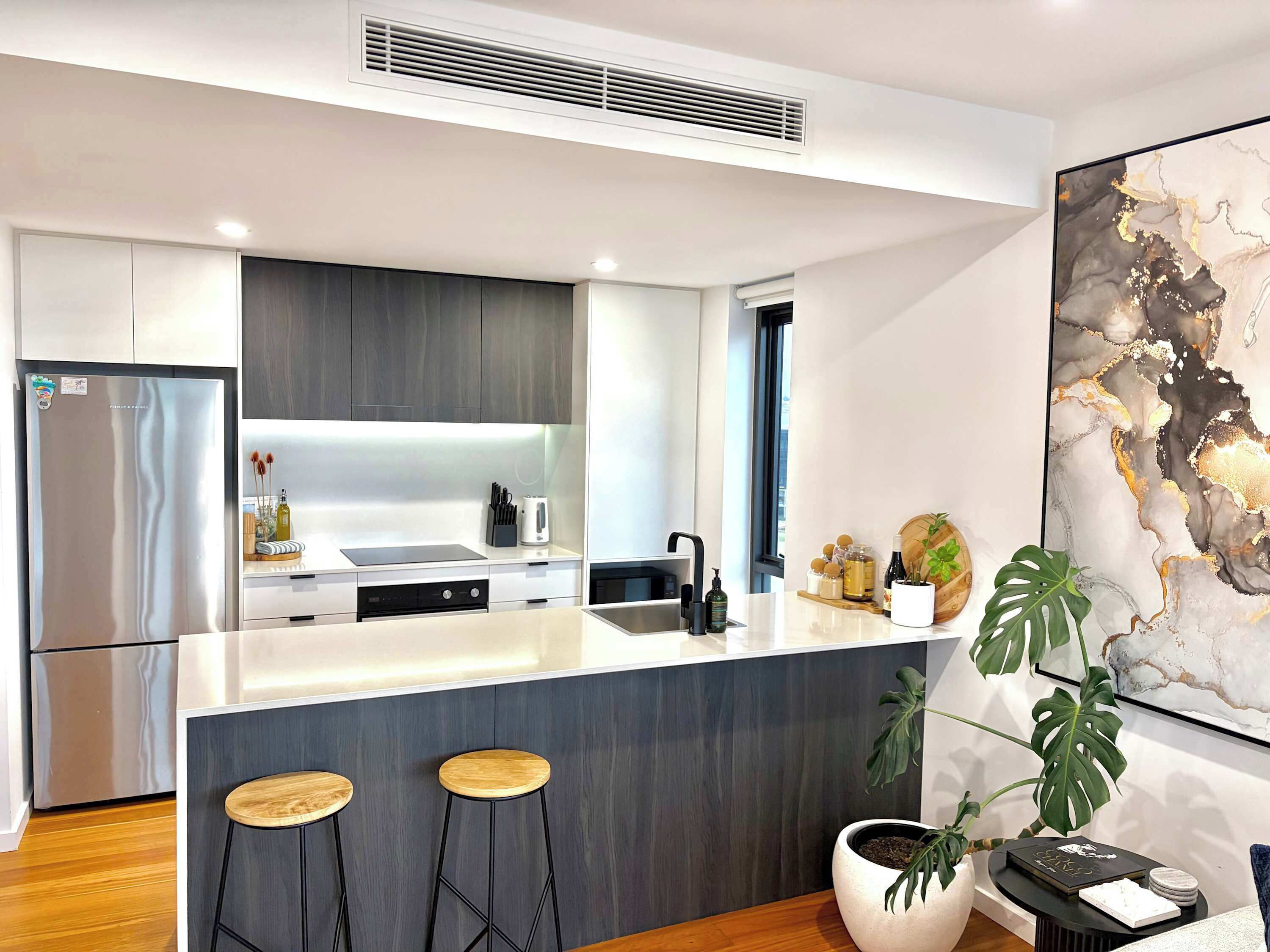 A modern kitchen with a central island, bar stools, a stainless steel refrigerator, and a large abstract painting on the wall.
