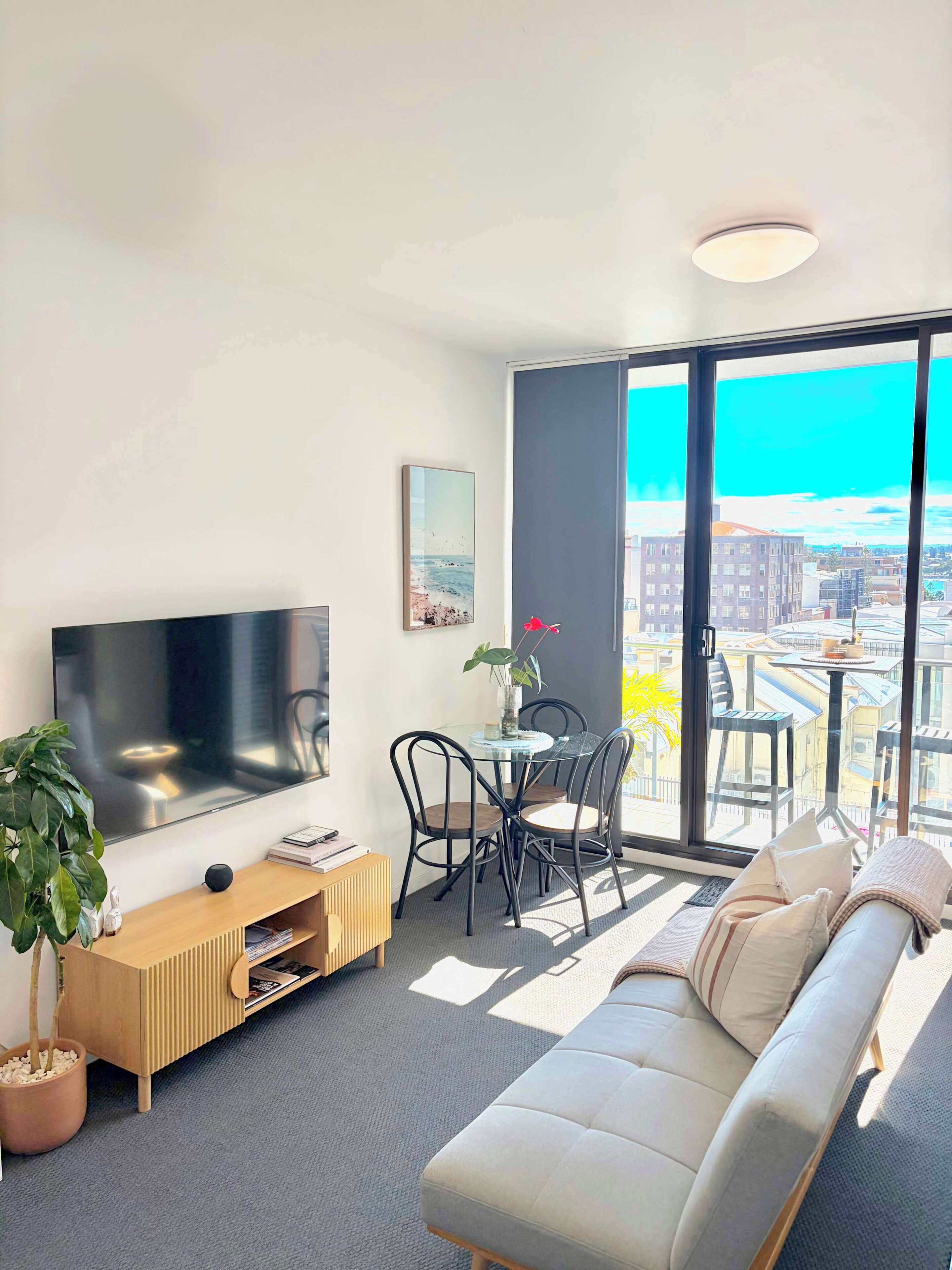 A modern living room with a flat-screen TV, a sofa, a dining table with chairs, and a large window offering a city view.