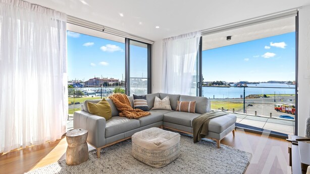 A modern living room with a grey sectional sofa, a round ottoman, and a view of a marina through large windows. - Newcastle Harbour, 2-Apartments 4-Bed 4-Bath 2-Car (Wickham)