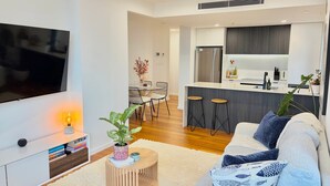 A modern living room with a flat-screen TV, a coffee table with a plant, a sofa with patterned cushions, and a kitchen area with a refrigerator and bar stools. - Newcastle Harbour, 2-Apartments 4-Bed 4-Bath 2-Car (Wickham)