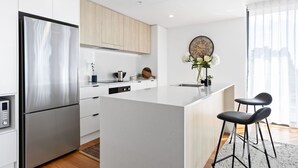A modern kitchen with a stainless steel refrigerator, wooden cabinets, a central island with bar stools, and a wall-mounted clock. - Newcastle Harbour, 2-Apartments 4-Bed 4-Bath 2-Car (Wickham)
