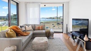 A modern living room with a large window, a sofa, a TV, and a view of a marina. - Newcastle Harbour, 2-Apartments 4-Bed 4-Bath 2-Car (Wickham)