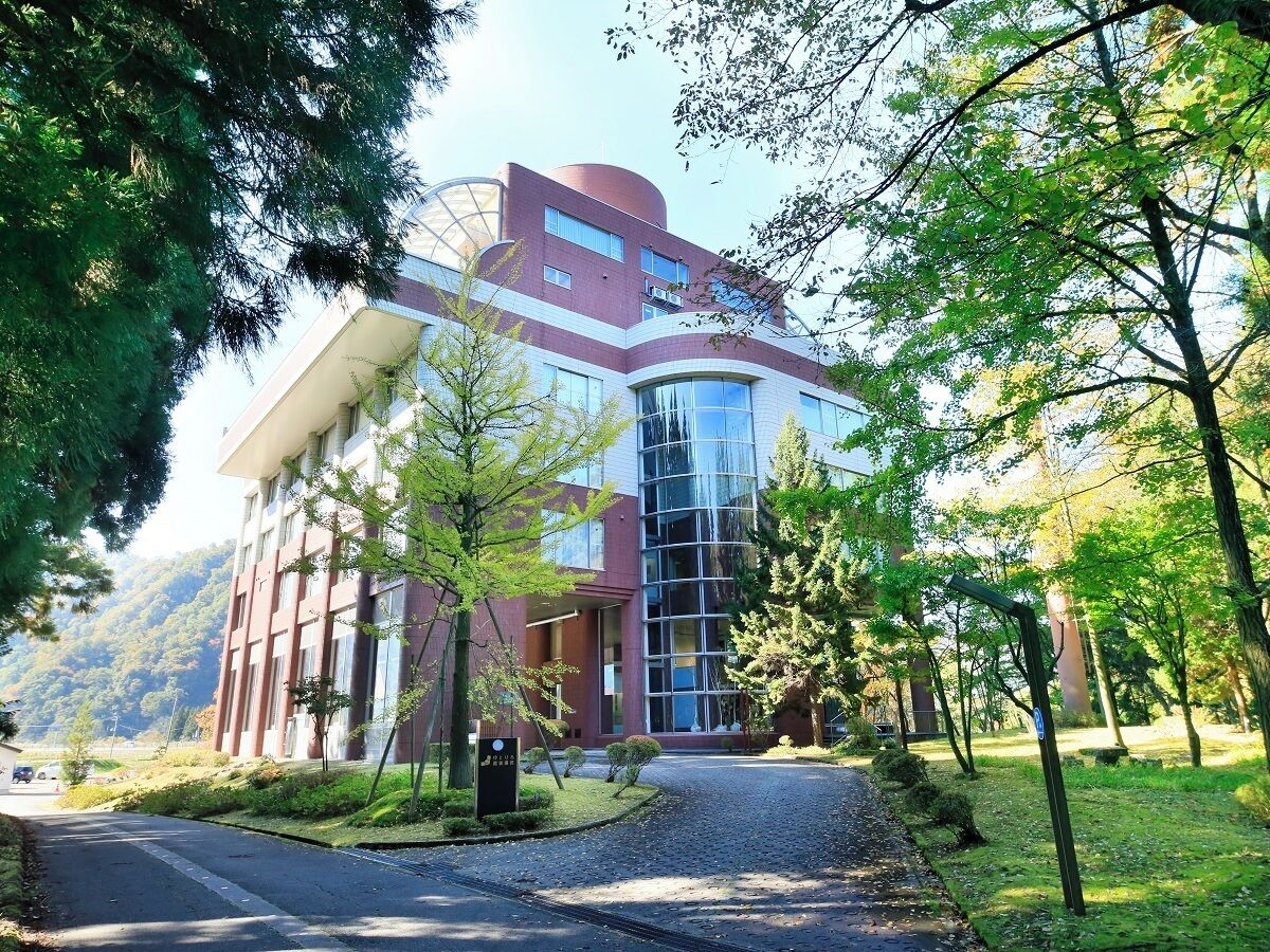 A modern building with a curved glass facade, surrounded by a well-maintained garden with trees and a gravel path leading to the entrance.