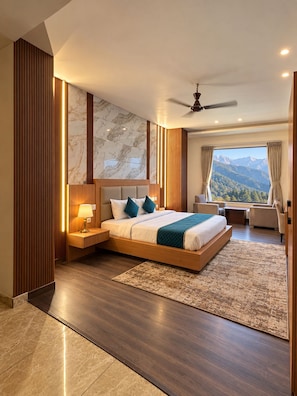 A modern bedroom with a large bed, a nightstand, a ceiling fan, and a view of mountains through the window. - Hotel vaikunth (Dharamshala)