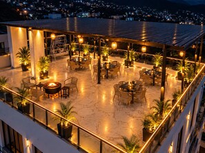 An outdoor patio area with multiple tables and chairs, surrounded by potted plants and illuminated by string lights. - Hotel vaikunth (Dharamshala)