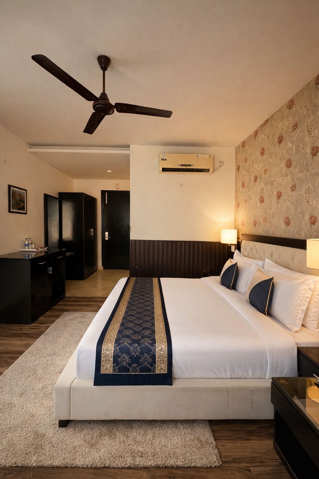 A hotel room with a bed, a ceiling fan, an air conditioning unit, a desk, and a wardrobe.
