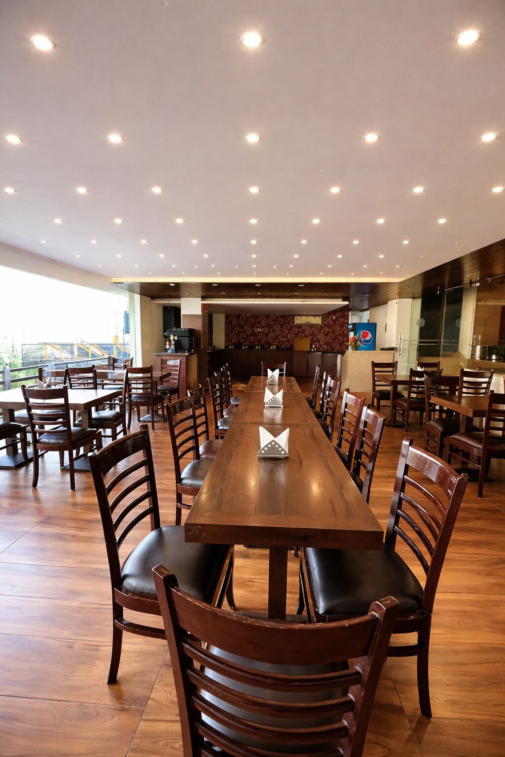 Een ruime eethoek met meerdere tafels en stoelen, een keuken op de achtergrond en een groot raam met uitzicht.