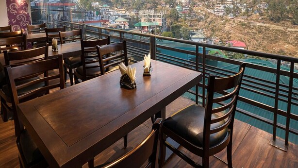 Un restaurante con mesas y sillas de madera, vistas a un paisaje montañoso y una barandilla con vista a una masa de agua.