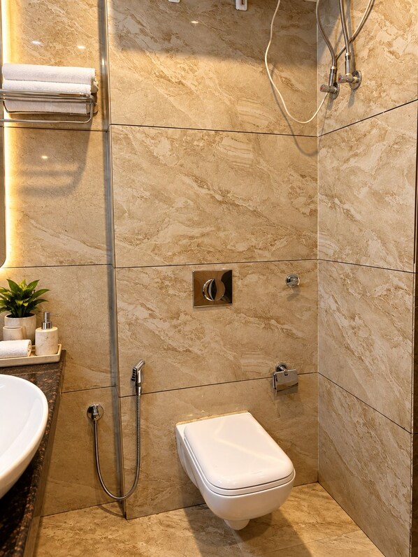 A bathroom with a marble wall, a white toilet, a shower with a handheld hose, and a towel rack with a towel. - Hotel vaikunth (Dharamshala)