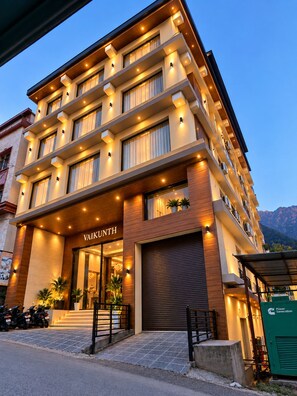 A multi-story hotel with a modern design, featuring a prominent entrance with a name sign, a covered outdoor area, and a view of the surrounding landscape. - Hotel vaikunth (Dharamshala)