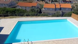 Ein rechteckiger Pool mit glasklar blauem Wasser, umgeben von einem Metallzaun, mit Blick auf eine Küstenstadt mit Häusern und einer Hügelkette im Hintergrund.