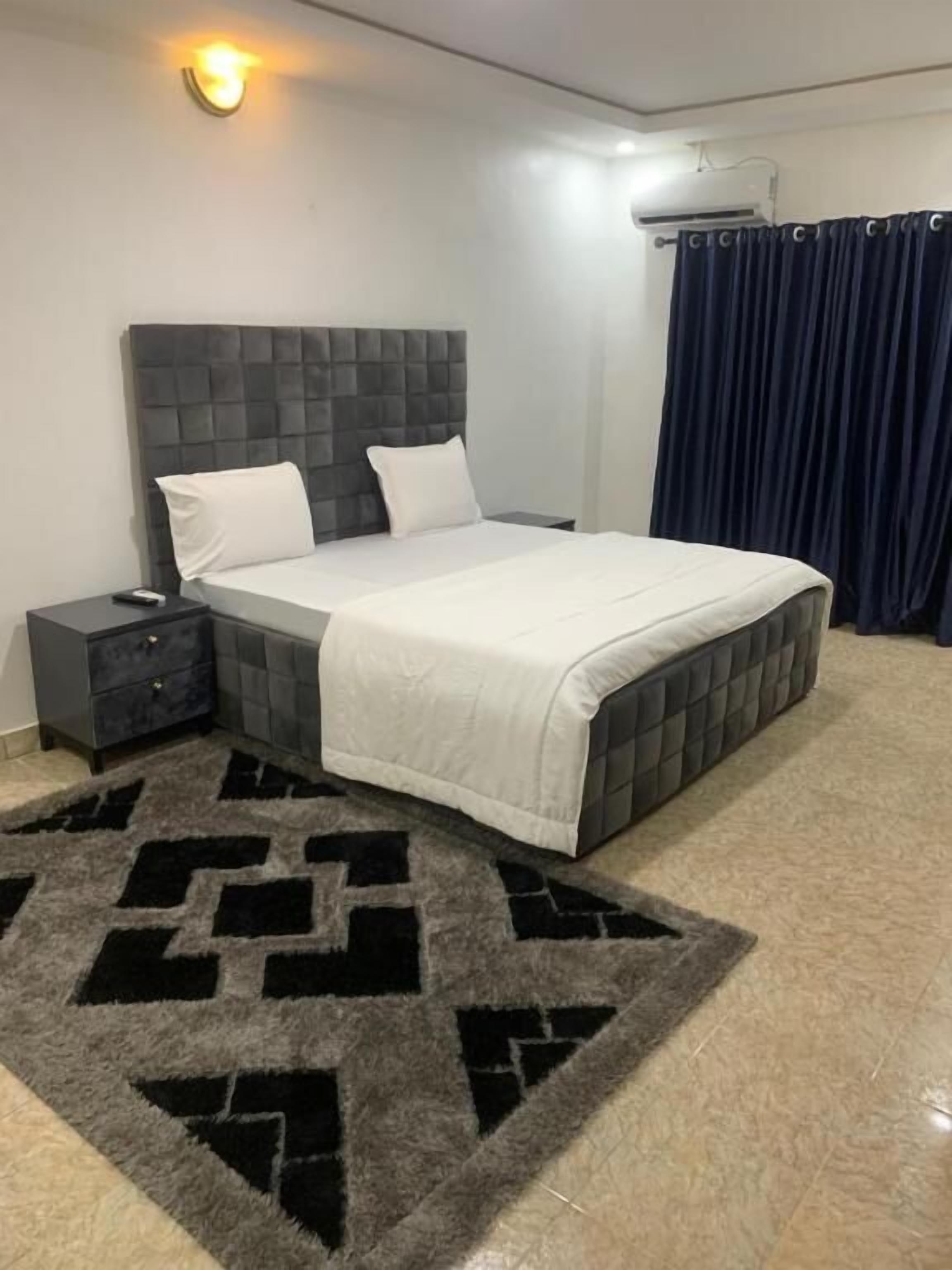 Une chambre à coucher avec une tête de lit capitonnée, un lit avec une literie blanche, une table de chevet, un tapis à motifs et un rideau foncé.